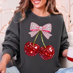 May include: Dark gray sweatshirt featuring a sparkling pink bow and two red cherries. The cherries have a faceted, gem-like appearance. The bow and cherries are the focal point of the design, creating a playful and eye-catching look.