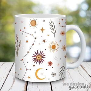 May include: White ceramic mug featuring a celestial design with suns, stars, flowers, and a crescent moon in shades of brown, orange, and gold. The mug has a white handle and the text "we design you create" at the bottom.