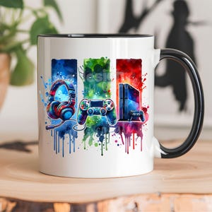May include: White ceramic mug with a black rim and handle. The mug features a colorful watercolor design of a video game controller, headphones, and a game console. The design is surrounded by colorful splatters of paint.
