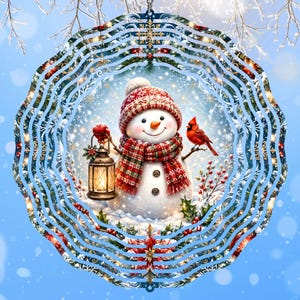 May include: A circular metal wind spinner featuring a central illustration of a snowman wearing a red patterned hat and scarf. The snowman holds a lit lantern, with a red cardinal perched on a branch nearby, surrounded by holly and snowflakes.