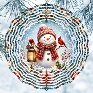 May include: A circular metal wind spinner featuring a festive winter scene with a snowman wearing a red patterned hat and scarf. The snowman holds a glowing lantern, accompanied by a red cardinal bird, holly, and poinsettia accents.