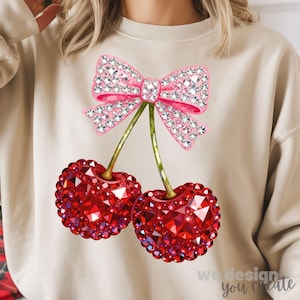 May include: Beige sweatshirt featuring a pink bow and two red cherries, all embellished with sparkling rhinestones. The cherries have green stems. The text "we design you create" is at the bottom right.