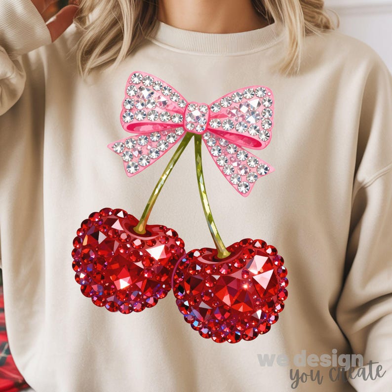 May include: Beige sweatshirt featuring a pink bow and two red cherries, all embellished with sparkling rhinestones. The cherries have green stems. The text "we design you create" is at the bottom right.