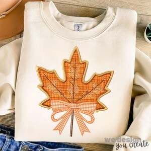 May include: Cream-coloured sweatshirt with a large, embroidered orange maple leaf with a plaid pattern and a gingham bow. The leaf has a gold outline and is centered on the front. The text "we design you create" is at the bottom right.