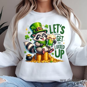 May include: White sweatshirt featuring a raccoon wearing a green hat and scarf, holding a green beer. The raccoon sits on a pile of gold coins with a pot of gold nearby. The text "LET'S GET LUCED UP" is in green.