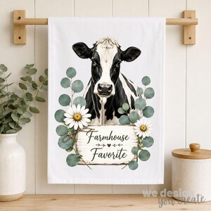 Può includere: Un canovaccio bianco con l'illustrazione di una mucca bianca e nera circondata da foglie di eucalipto verdi e due margherite bianche. Sotto la mucca, un cartello in legno riporta la scritta Farmhouse Favorite in caratteri neri.