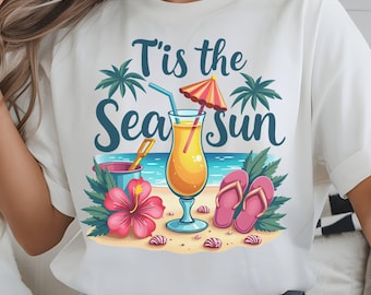 May include: White t-shirt featuring a beach scene graphic. The design includes a cocktail, pink flip-flops, a hibiscus flower, and the text "T'is the Sea Sun". Palm trees and a bucket with a shovel complete the summer scene.