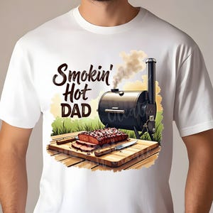 May include: White t-shirt with a graphic of a smoker with smoke coming out of it, a piece of meat on a cutting board, and the text "Smokin' Hot DAD".