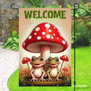 May include: A garden flag with a red and white mushroom with two green frogs sitting under it. The flag says "Welcome to our pad".