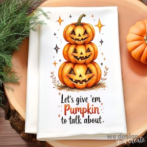 Puede incluir: Toalla de cocina blanca con temática de Halloween con una pila de tres calabazas naranjas talladas con caras sonrientes. La toalla tiene el texto "Let's give 'em Pumpkin to talk about". Estrellas y hojas decorativas rodean las calabazas.