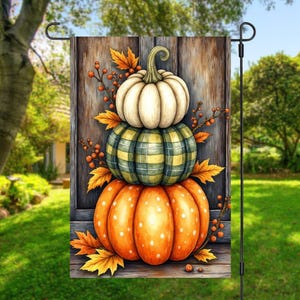 May include: A decorative garden flag featuring a stack of three pumpkins. The bottom pumpkin is orange with white spots, the middle one is green and yellow plaid, and the top is white. Autumn leaves and berries surround the pumpkins.