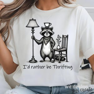 Puede incluir: Camiseta blanca con una ilustración en blanco y negro de un mapache sosteniendo una lámpara y una taza de té. El mapache lleva un pañuelo. El texto "I'd rather be Thrifting" está impreso debajo.