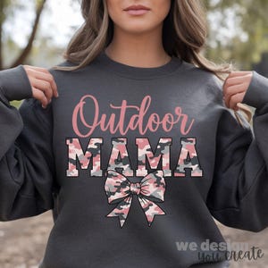 以下が含まれることがあります： ピンクと迷彩柄で「Outdoor MAMA」の文字が入った濃いグレーのスウェットシャツ。テキストの下には迷彩柄のリボンがあります。スウェットシャツはカジュアルなスタイルです。