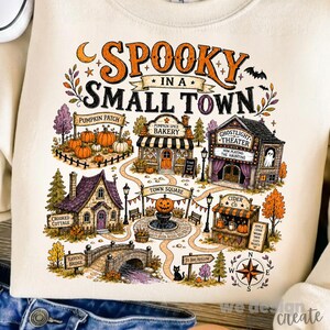 May include: A cream-colored sweatshirt featuring a colorful graphic of a small town with the text SPOOKY IN A SMALL TOWN. The illustration includes a pumpkin patch, bakery, theater, cottage, cider stand, bridge, and a compass rose in autumn colors.