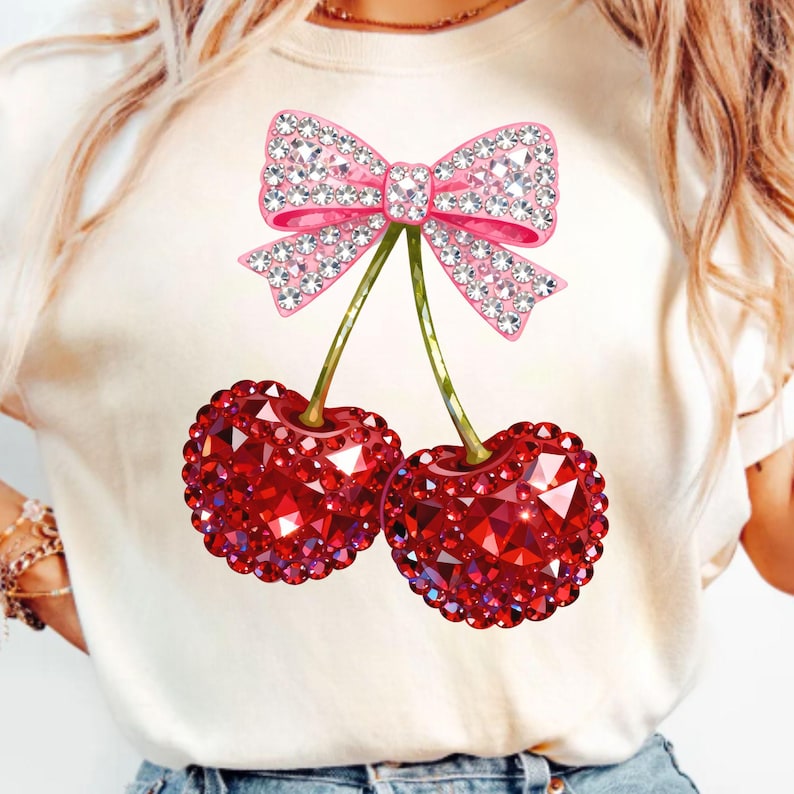 May include: Cream-colored t-shirt featuring a sparkling pink bow and two red cherries. The cherries and bow are embellished with rhinestones. The cherries have green stems. The shirt is a casual top.