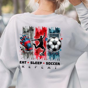 May include: White sweatshirt with a watercolor design of a soccer player, a soccer ball, and a goal. The text "Eat Sleep Soccer Repeat" is printed below the design.