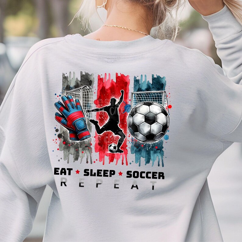 May include: White sweatshirt with a watercolor design of a soccer player, a soccer ball, and a goal. The text "Eat Sleep Soccer Repeat" is printed below the design.