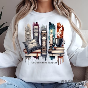 May include: White sweatshirt featuring a watercolour design of stacked books, a lit candle, and a cup of coffee. The text "Just one more chapter" is printed below the design.