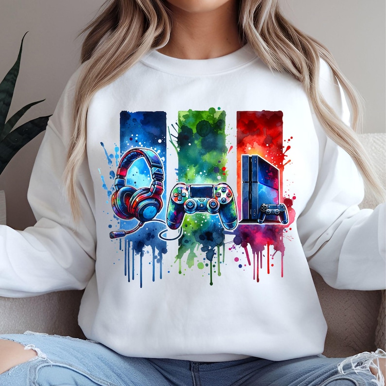 May include: White sweatshirt with a colorful watercolor design of a headset, video game controller, and a video game console. The design is surrounded by colorful paint splatters.
