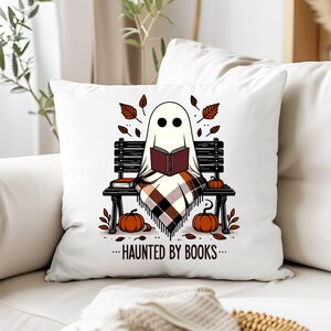 以下が含まれることがあります： ハロウィーンをテーマにしたデザインの白い正方形のクッション。幽霊が本を読んでいます。カボチャと秋の葉に囲まれたベンチに座っています。「...HAUNTED BY BOOKS...」というテキストが下にあります。