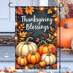 Könnte beinhalten: Eine Gartenflagge mit Thanksgiving-Motiv. Die Flagge zeigt die Worte "Thanksgiving Blessings" über einer Sammlung von Kürbissen in Orange und Weiß. Herbstblätter und Lichterketten schmücken den Hintergrund.