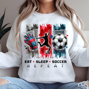 May include: White sweatshirt featuring a watercolour design of a football player, a football, and a pair of goalkeeper gloves. The text "Eat Sleep Soccer Repeat" is printed below the design.