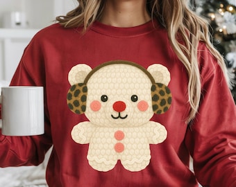 May include: A red sweatshirt with a cream teddy bear design. The bear has brown earmuffs with a leopard print, a red nose, and pink cheeks. The text "we design & create" is at the bottom right.