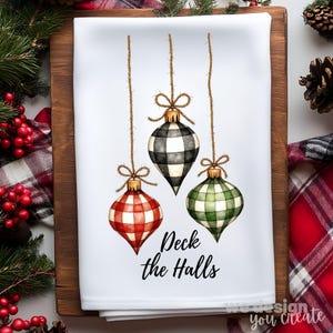 Puede incluir: Paño de cocina blanco con temática navideña con tres adornos colgantes a cuadros rojos, blancos y negros y verdes. El texto "Deck the Halls" está escrito en escritura negra debajo. El paño se muestra sobre una superficie de madera.