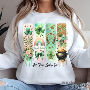 Puede incluir: Sudadera blanca con un diseño gráfico de cuatro rayas verticales con tréboles, un arcoíris, una herradura y una olla de oro. Las rayas están goteando pintura dorada. El texto "Get Your Lucky On" está impreso debajo del gráfico.