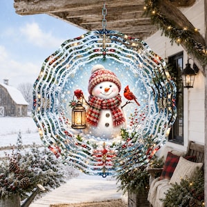 May include: A circular metal wind spinner featuring a central illustration of a snowman wearing a red plaid scarf and hat, holding a lit lantern, with a red cardinal perched nearby, surrounded by a snowy winter landscape and decorative snowflakes.