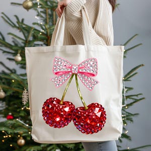 May include: Cream-colored tote bag featuring a pink bow and two red cherry designs. The bow and cherries are embellished with sparkling details. The bag is held up in front of a Christmas tree.
