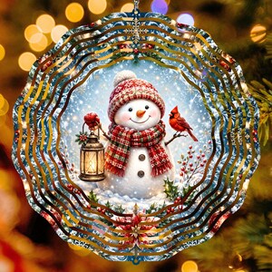 May include: A circular metal wind spinner featuring a snowman wearing a red patterned hat and scarf, holding a lit lantern. A red cardinal sits on a branch next to the snowman. The design includes holly, berries, and snowflakes on a blue background.