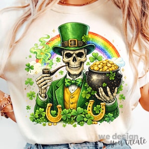 Op de afbeelding: Crèmekleurig T-shirt met een skelet verkleed als een leprechaun, compleet met een groene hoge hoed, jas en vlinderdas. Het skelet houdt een pijp en een pot goud vast, met een regenboog en klavers op de achtergrond. Gouden hoefijzers en de tekst "we design you create" zijn ook zichtbaar.