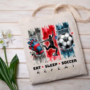 May include: A white canvas tote bag with a watercolor design of a soccer player, a soccer ball, and a goal. The text "Eat Sleep Soccer Repeat" is printed on the bag.