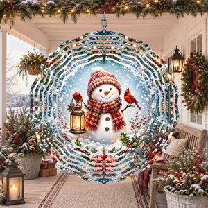 May include: A circular metal wind spinner featuring a central illustration of a snowman wearing a red patterned hat and scarf, holding a lit lantern, with a red cardinal perched on its arm, surrounded by a blue snowy background and holly.