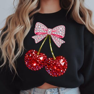 May include: Black sweatshirt featuring a pink bow with crystal embellishments and two red crystal-covered cherries. The cherries have green stems. The design is centered on the front of the sweatshirt.