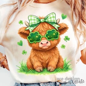 May include: A light-colored t-shirt featuring a cartoon Highland cow with a green bow, shamrock glasses, and shamrock accents. The cow is sitting in green grass. The text "we design you create" is at the bottom.