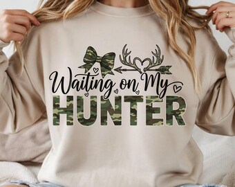 Op de afbeelding: Beige sweatshirt met de tekst "Waiting on My Hunter" in zwart schrift en camouflage letters. Het ontwerp bevat een strik, geweien, een pijl en harten. De sweatshirt heeft een neutrale kleur.