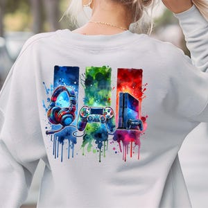 May include: A white sweatshirt with a colorful watercolor design of a video game controller, headphones, and a game console. The design is surrounded by colorful paint splatters.