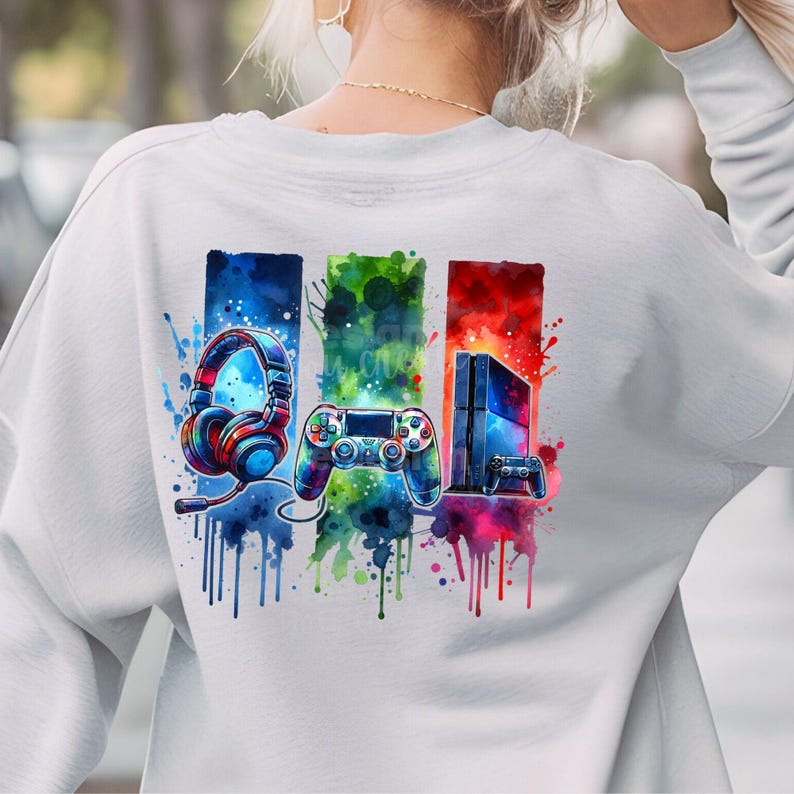May include: A white sweatshirt with a colorful watercolor design of a video game controller, headphones, and a game console. The design is surrounded by colorful paint splatters.