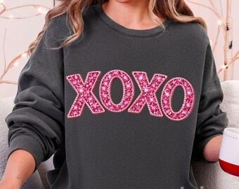 May include: Dark gray sweatshirt with the word "XOXO" in large, sparkling pink letters across the front. The sweatshirt is a relaxed fit and has long sleeves. The text "we design create" is at the bottom right.