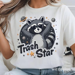 Puede incluir: Camiseta blanca con un mapache de dibujos animados con gafas de sol y una pose relajada. El mapache está rodeado de estrellas y planetas, con el texto "Trash Star" debajo.