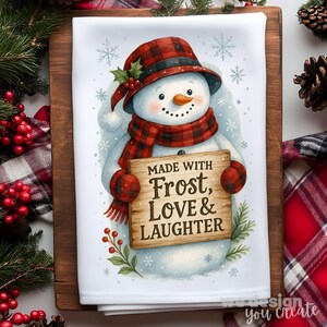 May include: A white kitchen towel featuring a snowman illustration. The snowman wears a red plaid hat, scarf, and mittens, and holds a wooden sign that reads "Made with Frost, Love & Laughter". Snowflakes and holly sprigs decorate the background.