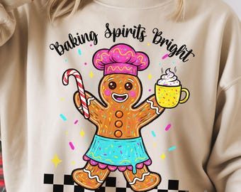 May include: A beige sweatshirt featuring a gingerbread person wearing a chef's hat and apron, holding a candy cane and a mug of whipped cream. The text "Baking Spirits Bright" is arched above the gingerbread man. Colorful sprinkles and stars decorate the design.