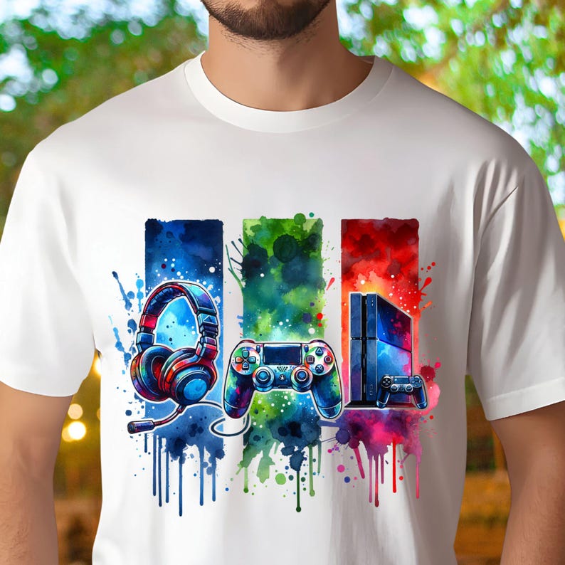 May include: A white t-shirt with a colorful watercolor design featuring a video game controller, headphones, and a game console. The design is in shades of blue, green, red, and purple.