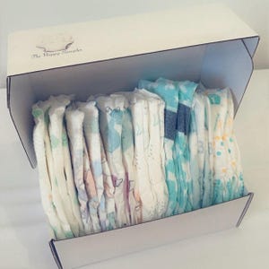 May include: A white box with the text "The Happy Sampler" on the top. The box is open and filled with a variety of baby nappies with different patterns and colours.