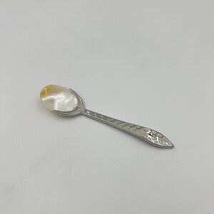 以下が含まれることがあります： 白と黄色の虹色に輝くマザーオブパールのボウルと、銀色の金属製ハンドルを備えた小さなスプーン。ハンドルは全長にわたってハンマーで叩いたような質感の仕上げが施されており、先端は金属光沢のある尖った形状になっています。