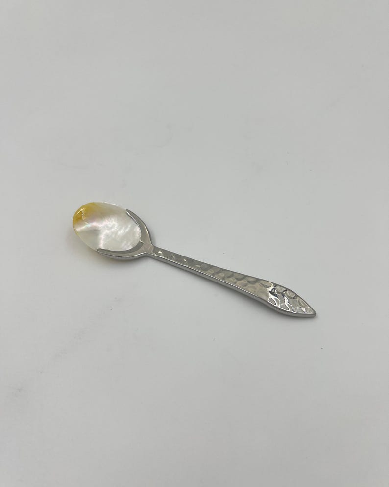 以下が含まれることがあります： 白と黄色の虹色に輝くマザーオブパールのボウルと、銀色の金属製ハンドルを備えた小さなスプーン。ハンドルは全長にわたってハンマーで叩いたような質感の仕上げが施されており、先端は金属光沢のある尖った形状になっています。