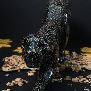 May include: A black ceramic panther figurine with a spotted pattern. The figurine is posed in a walking position with its head turned to the side. The figurine is sitting on a bed of wood shavings.