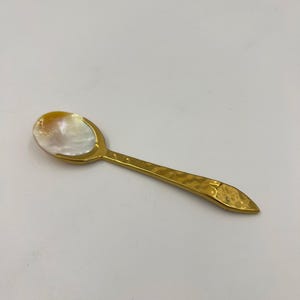 以下が含まれることがあります： 虹色に輝く白いマザーオブパールのボウルと、ハンマーで叩いたような質感のゴールドトーンの金属製ハンドルを備えた小さなスプーン。ハンドルは先端に向かって細くなっており、ボウルは金属製のフレームにしっかりと固定されています。